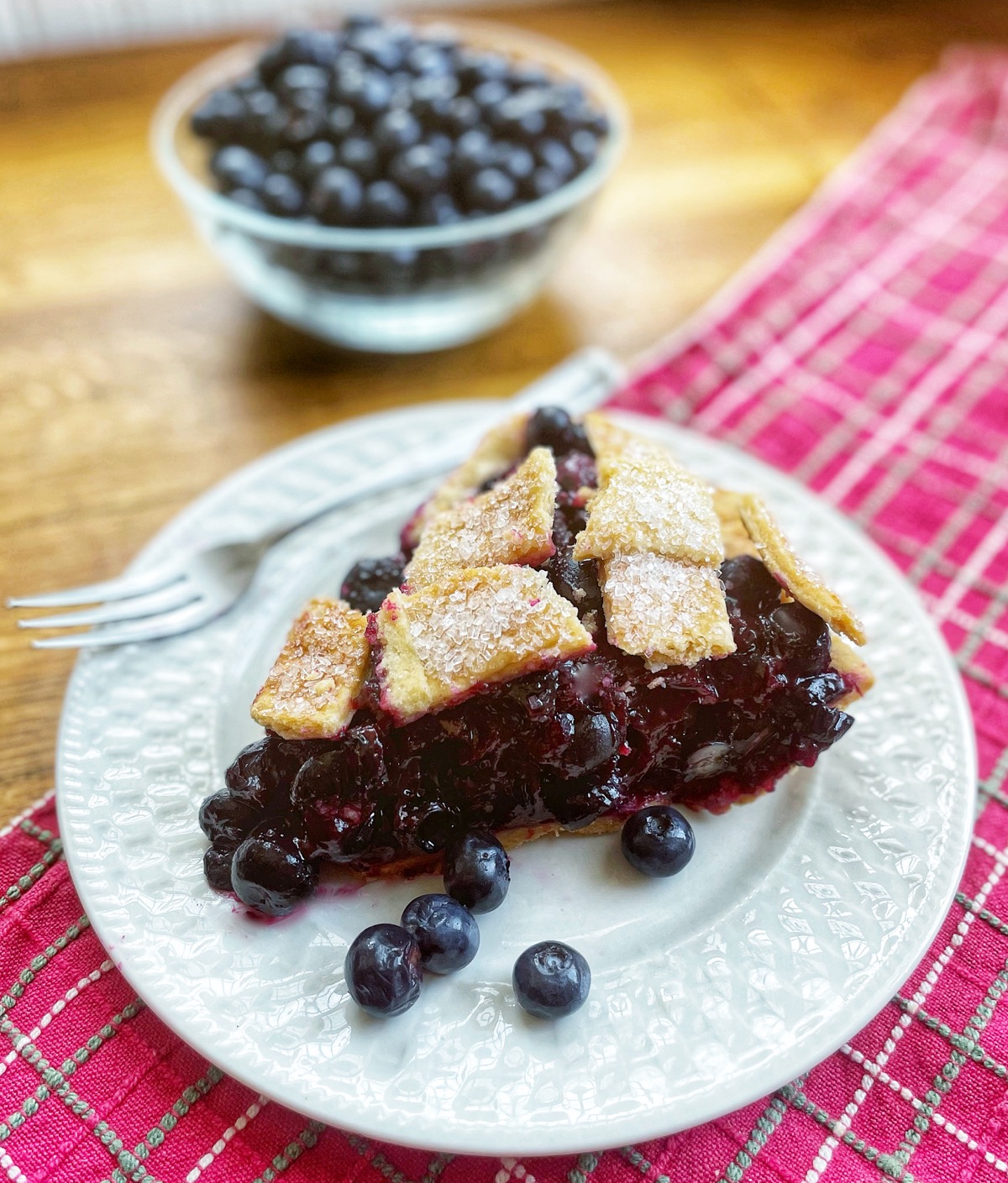 Slice of blueberry pie on a plate, bowl of blueberries in the background.