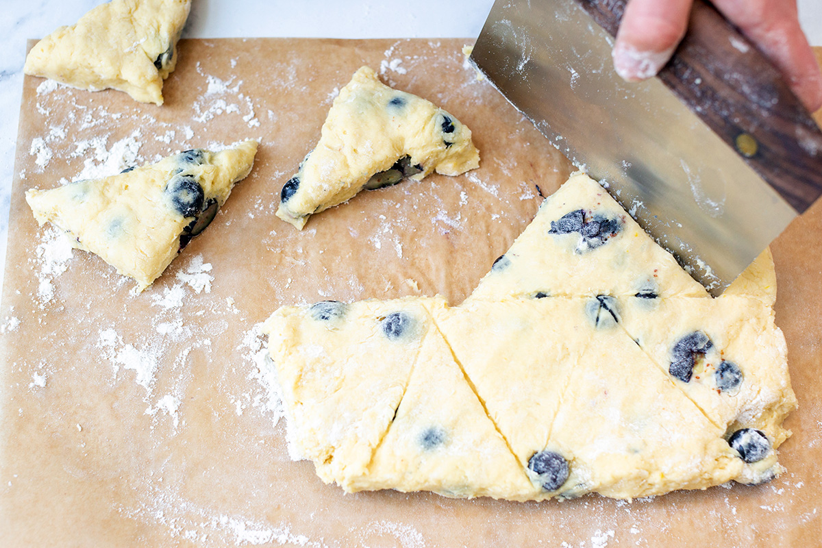 A baker using a bench knife to cut fresh blueberry scone dough into 10 triangle pieces