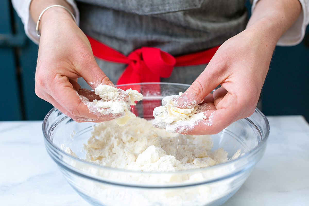 A baker's hands working 1/2" chunks of cold butter into the dry ingredients for scones in a bowl