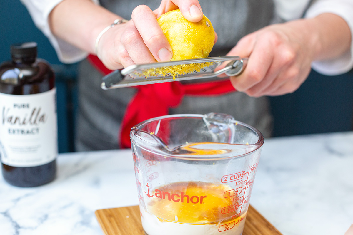 A baker zesting a lemon over a measuring cup of eggs, milk, and vanilla extract