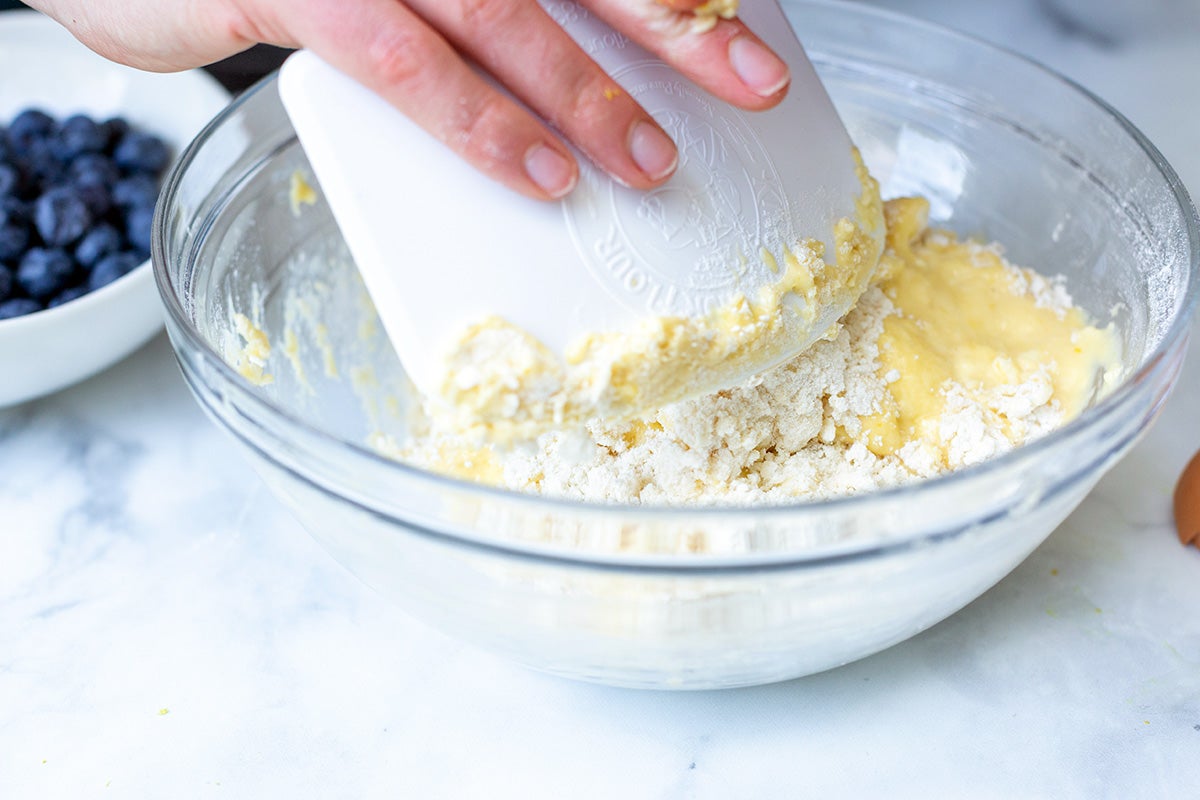 A baker using a dough scraper to fold the wet and dry ingredients for fresh blueberry scones together in a mixing bowl