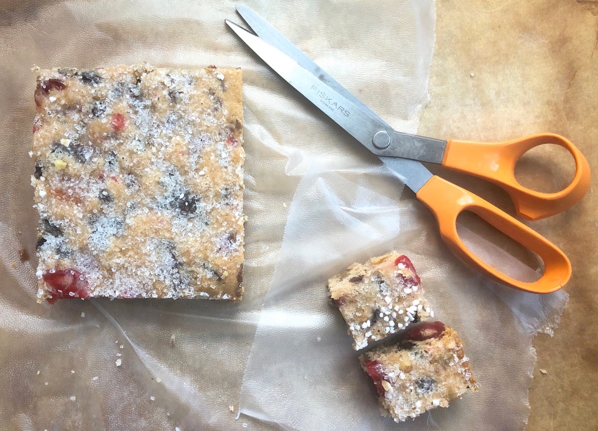 Large square and smaller squares of fruitcake bars on pieces of plastic wrap, with a pair of scissors.