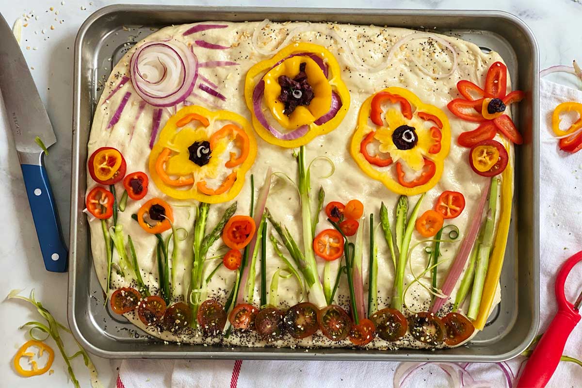 A unbaked focaccia topped with vegetables in a garden scene