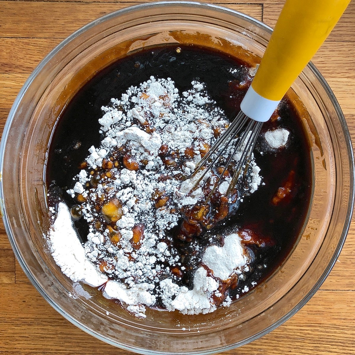 Liquid ingredients for gingerbread being poured atop dry ingredients in a glass bowl.