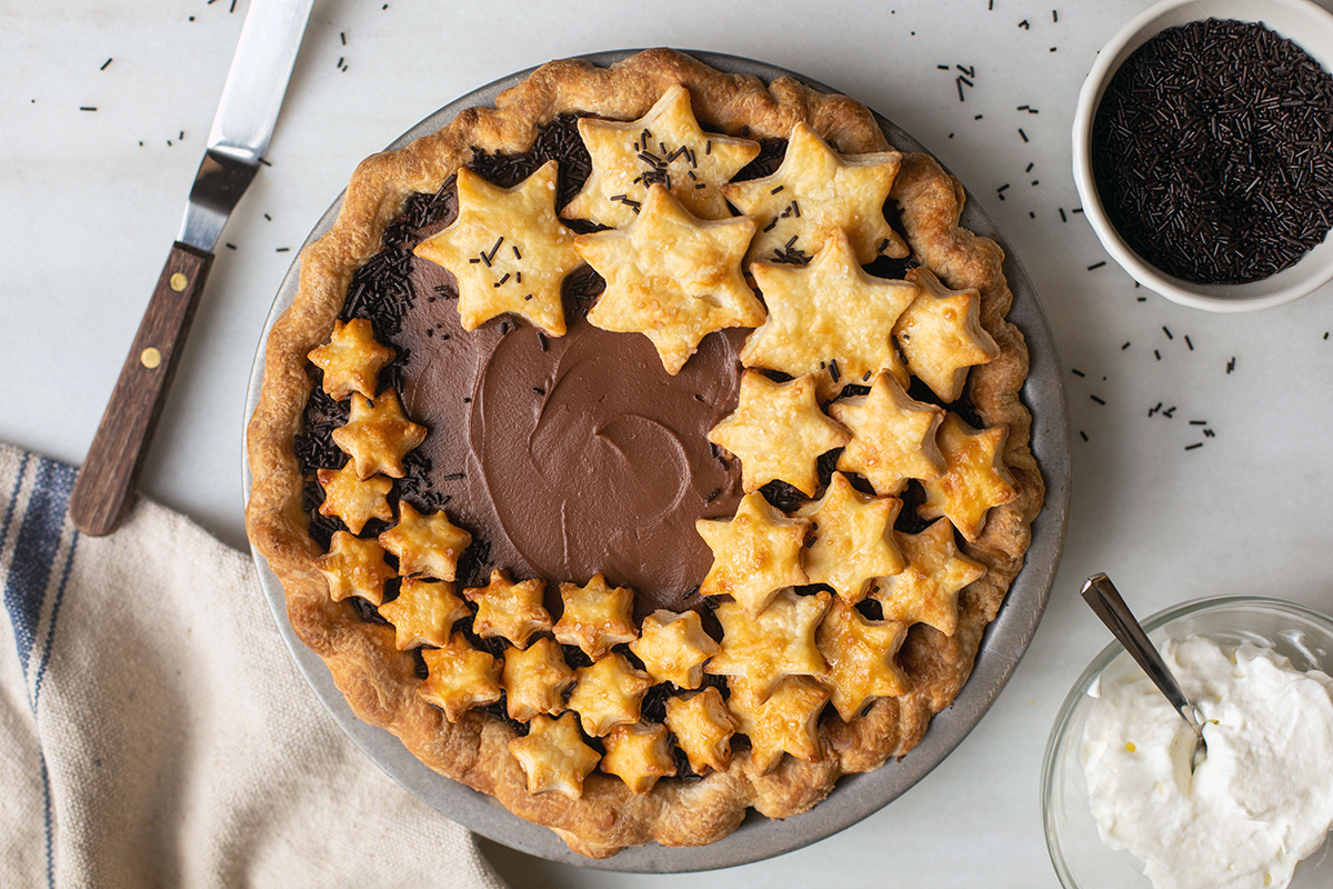 A chocolate cream pie topped with star-shaped cutouts next to a bowl of chocolate sprinkles