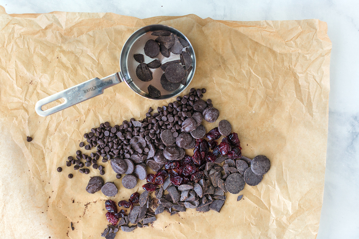 Chopped chocolate, dried cranberries, and chocolate chips on a piece of parchment paper with a measuring cup