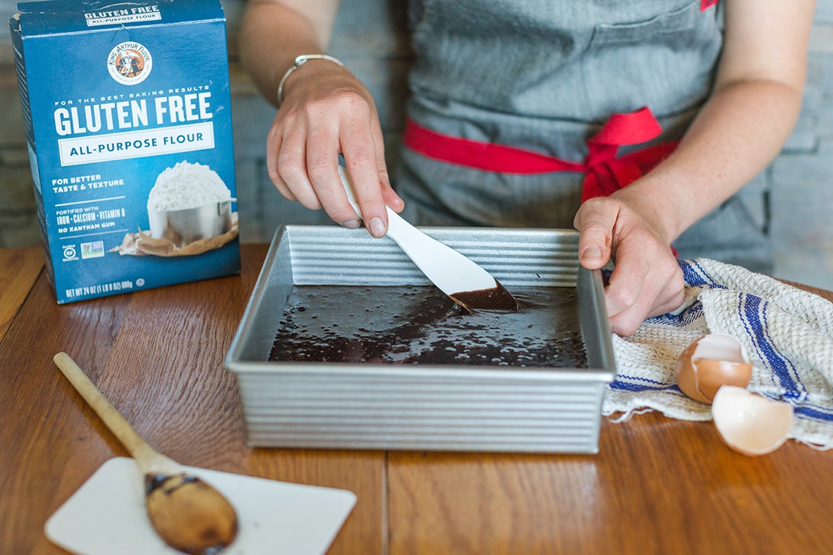 A baker's hand spreading gluten-free brownie batter into the edges of a square pan before baking