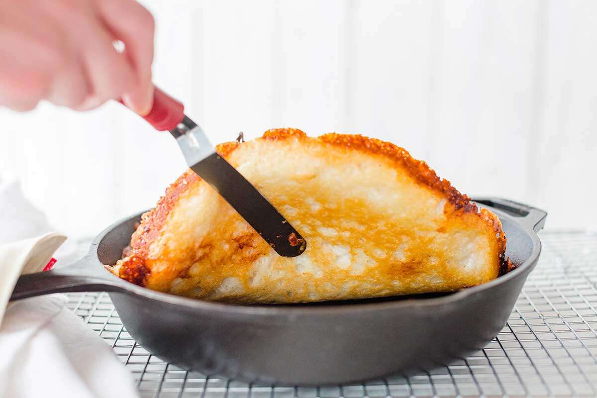 A baker using a spatula to check the underside of a cast iron pan pizza to make sure it's golden brown