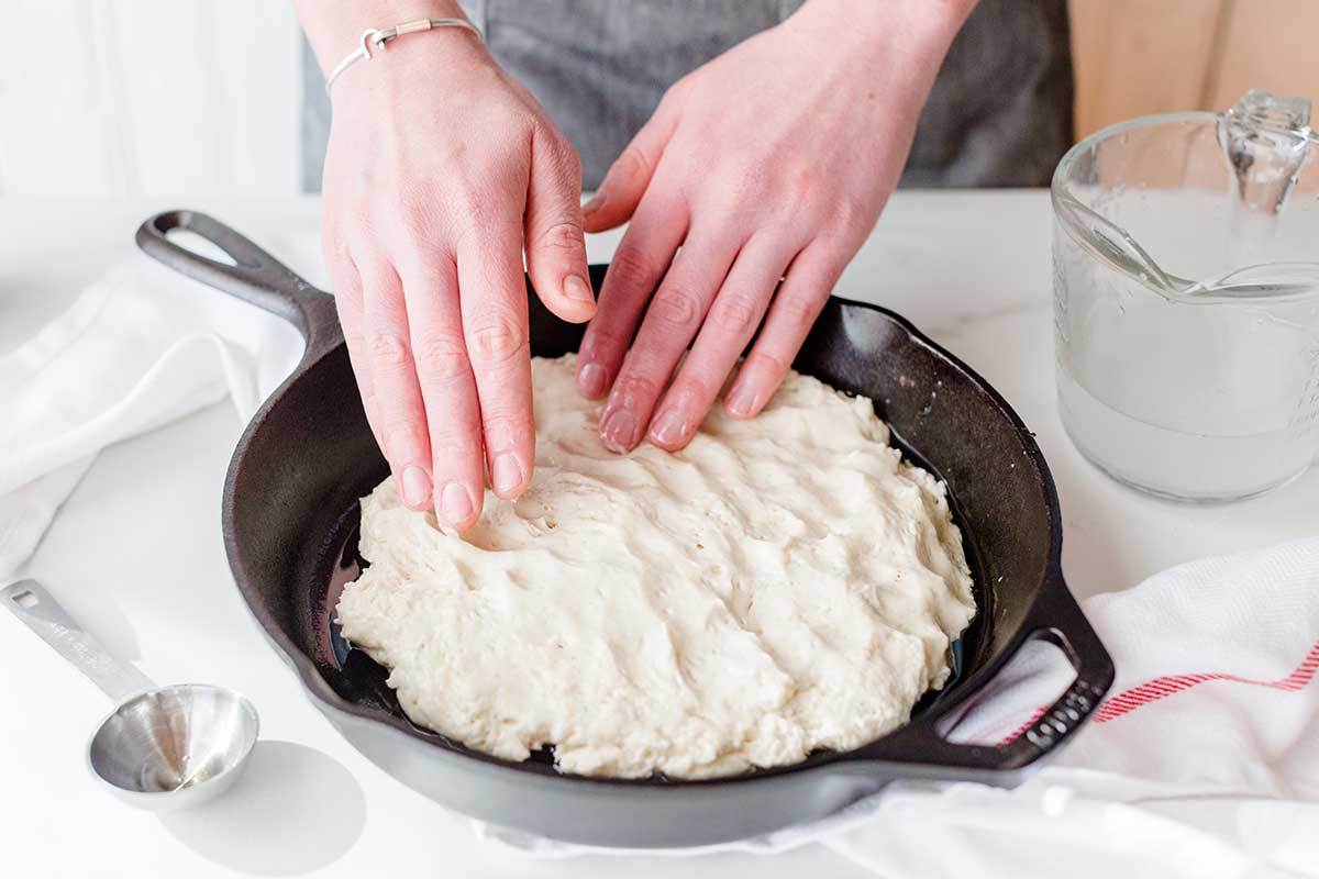 A baker using her hands to smooth out gluten-free pizza dough to the edges of a cast iron pan