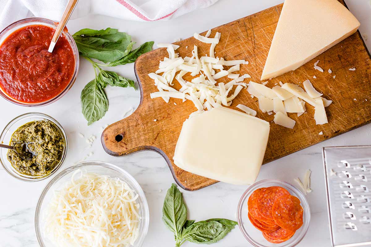 A kitchen table set up with assorted cheeses, sauces, and pizza toppings