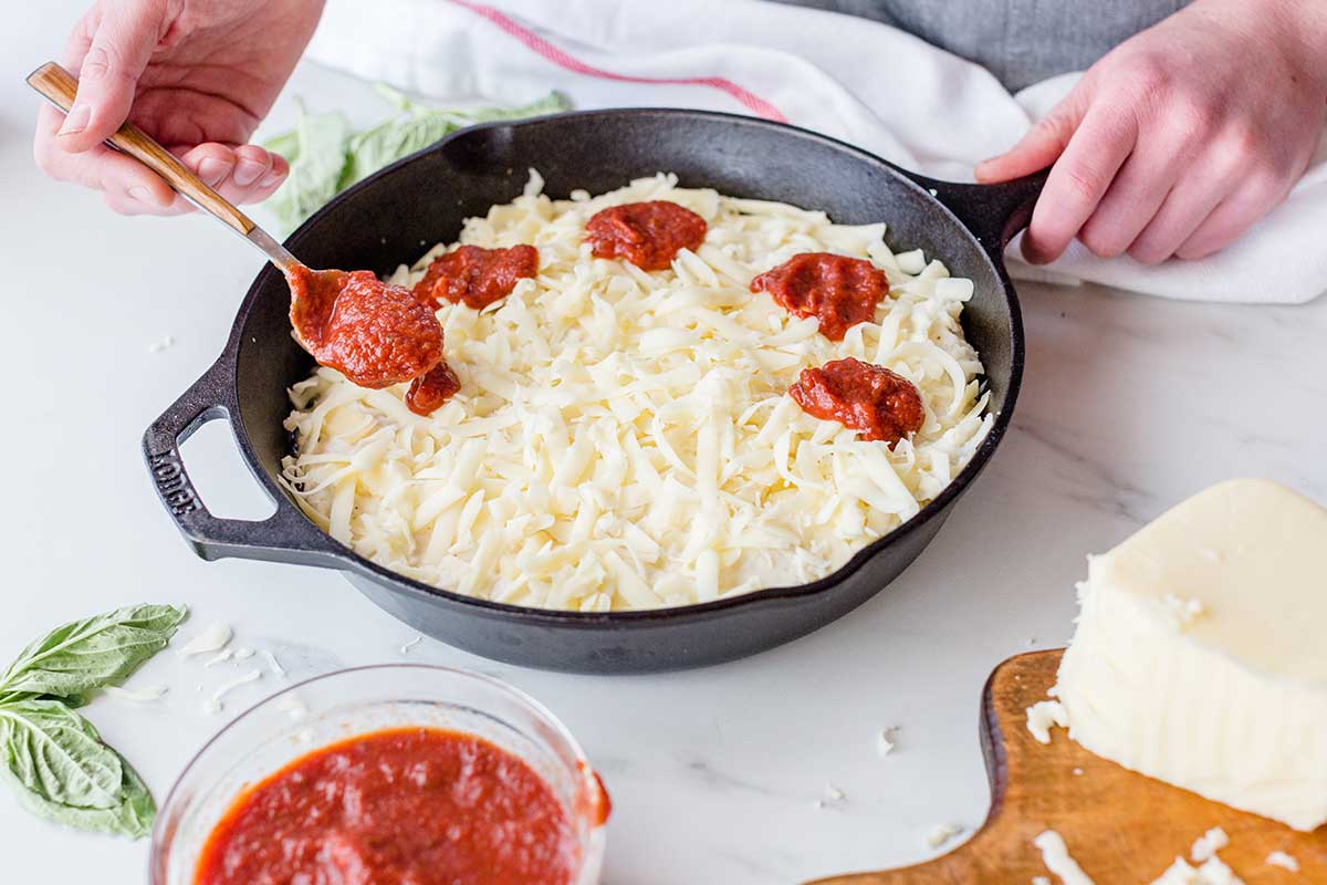 A baker dolloping tomato sauce over pizza dough topped with cheese