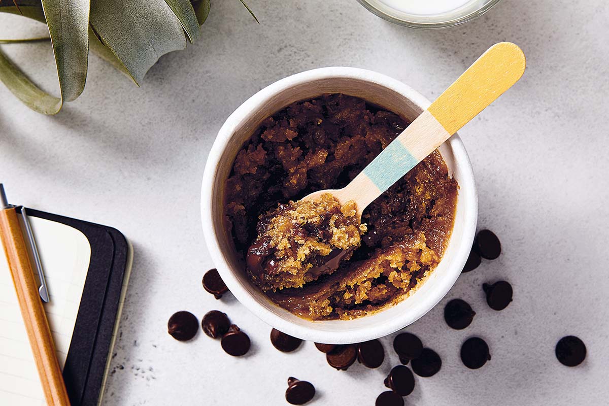 A cup of gluten-free single-serve chocolate chip cookie with a spoon and scattered chocolate chips on a kitchen table