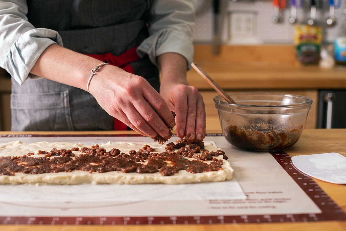 A baker’s hands sprinkling cinnamon filling over rolled out cinnamon roll dough