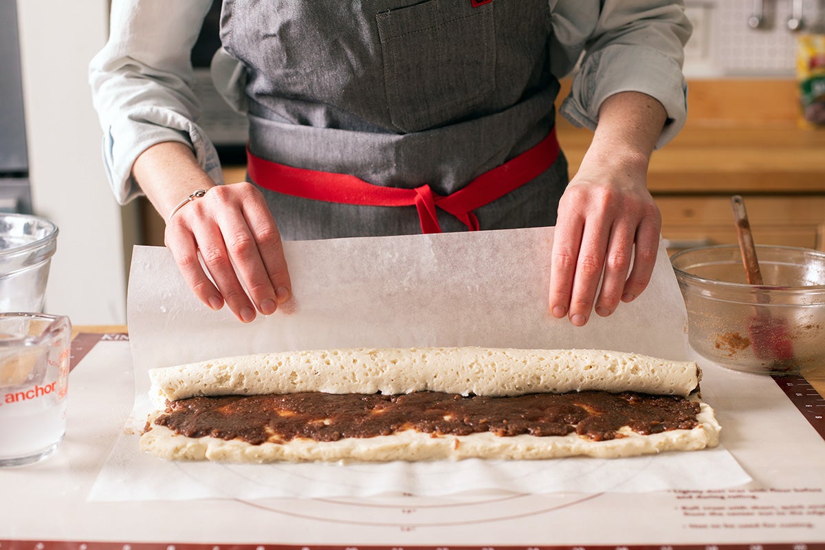 A baker rolling up gluten-free cinnamon roll dough using a piece of parchment paper to help lift it