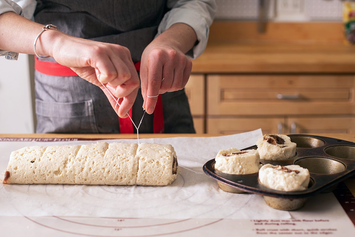 A baker using dental floss to slice a log of cinnamon roll dough into 8 pieces