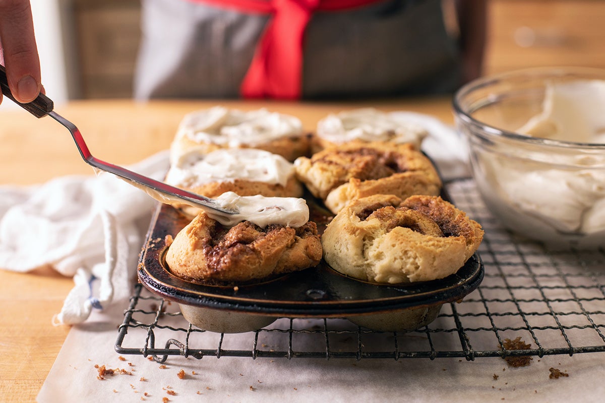 A baker icing gluten-free cinnamon rolls with cream cheese frosting using an offset spatula