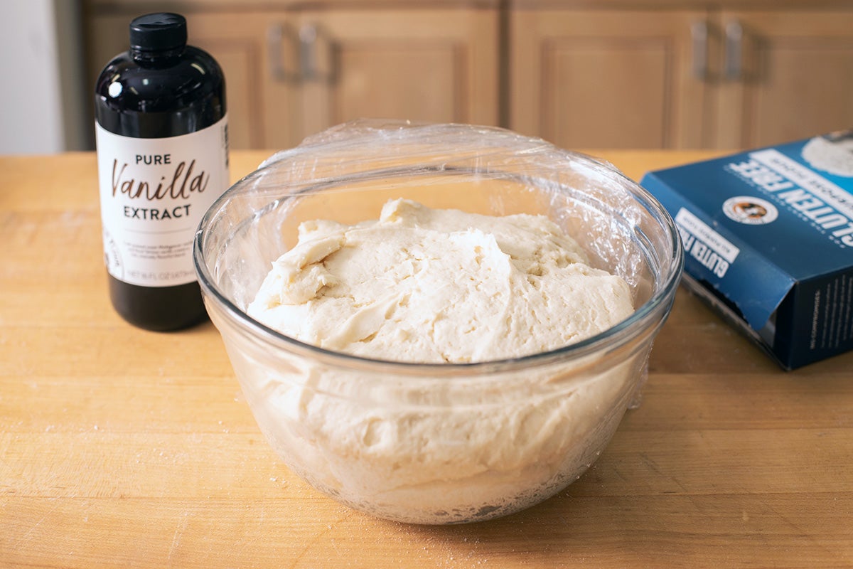 Gluten-free cinnamon roll dough resting in a glass bowl, covered with plastic, looking puffy and fully risen