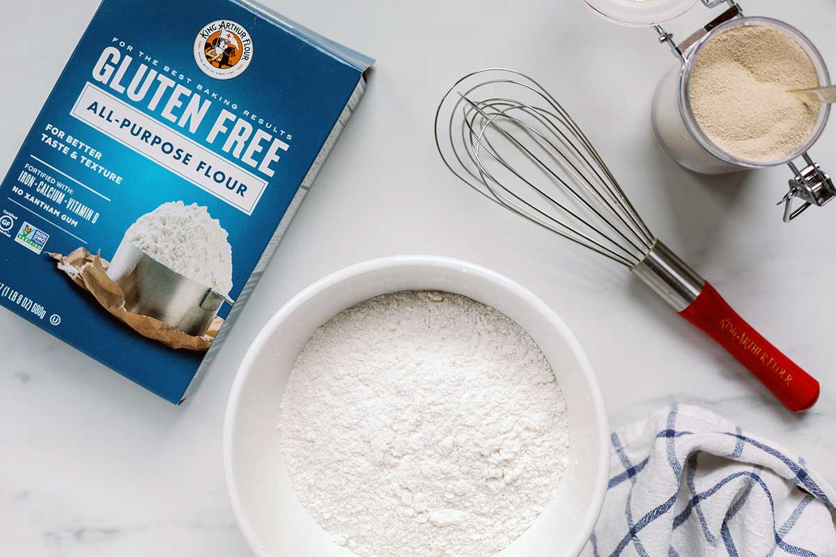 A bowl of King Arthur Gluten-Free Flour next to a whisk and a container of yeast