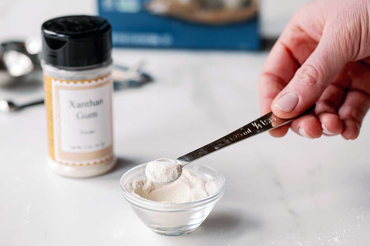 A baker measuring 1/4 teaspoon of xanthan gum from a small bowl