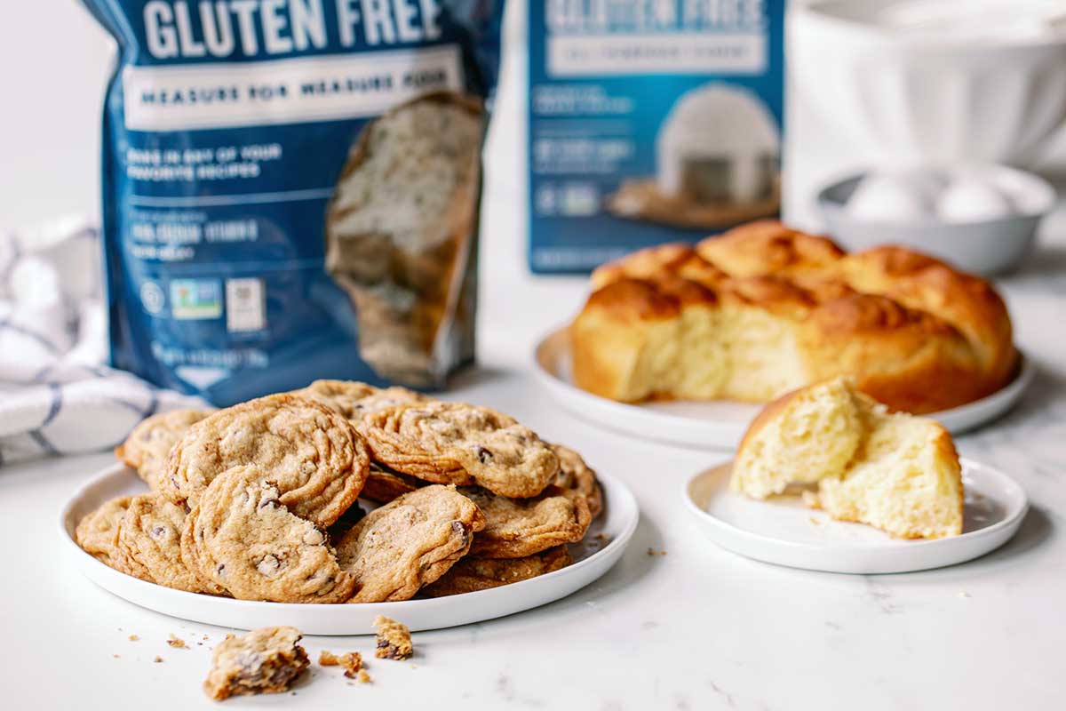A bag of Gluten-Free Measure for Measure Flour in front of a plate of chocolate chip cookies with gluten-free rolls in the background