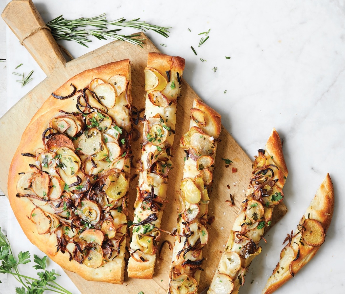 Potato focaccia garnished with fresh rosemary and charred onions on a cutting board.