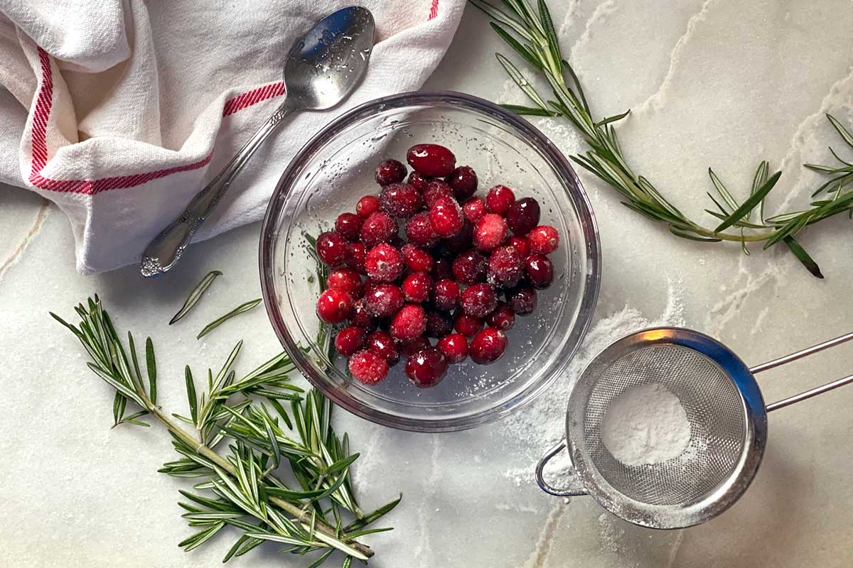 Sugared cranberries next to a few sprigs of rosemary and snow white non-melting sugar