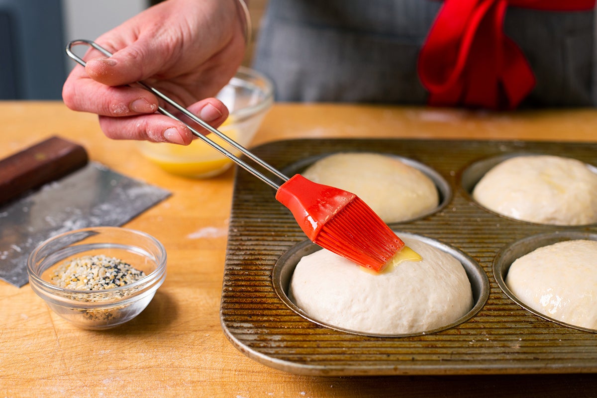 een bakker die volledig gestegen hamburgerbroodjes met een egg wash borstelt voordat hij in de oven gaat