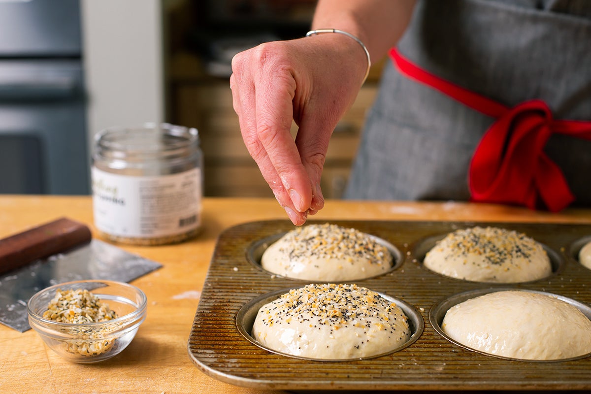 A baker sprinkling Everything Bagel Topping onto homemade burger buns before baking