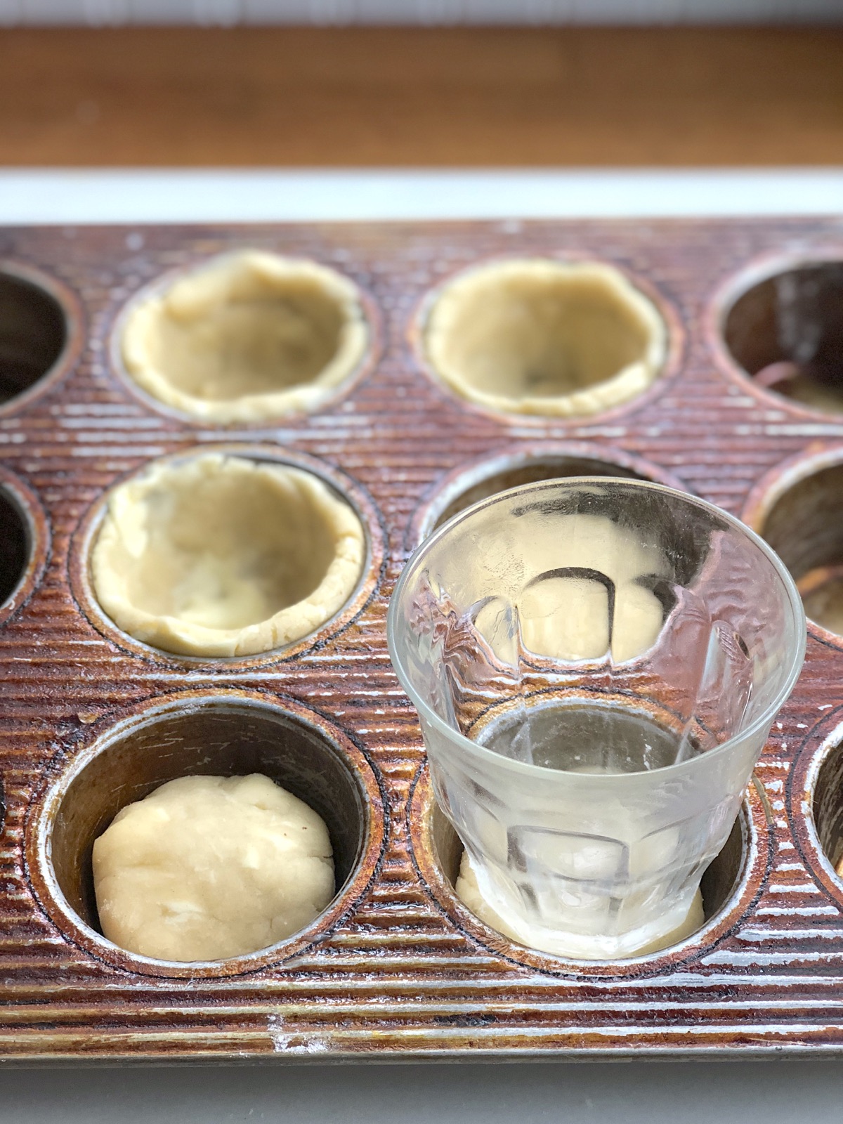Mini pie crusts being pressed into the wells of a muffin pan using a drinking glass.