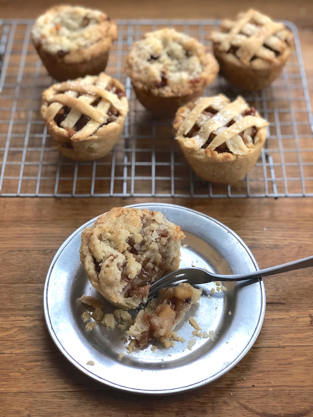 Apple mini pie on a plate with a fork, cut open, more mini pies in the background on a cooling rack.