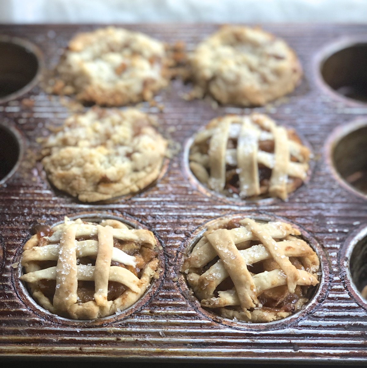 Mini apple pies baked in a mufin tin, some with lattice top crusts, some with crumb top crusts.