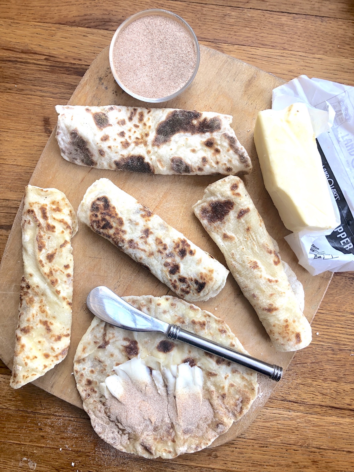 Lefse rolled into cylinders on a cutting board, one open being spread with butter and sprinkled with cinnamon sugar.