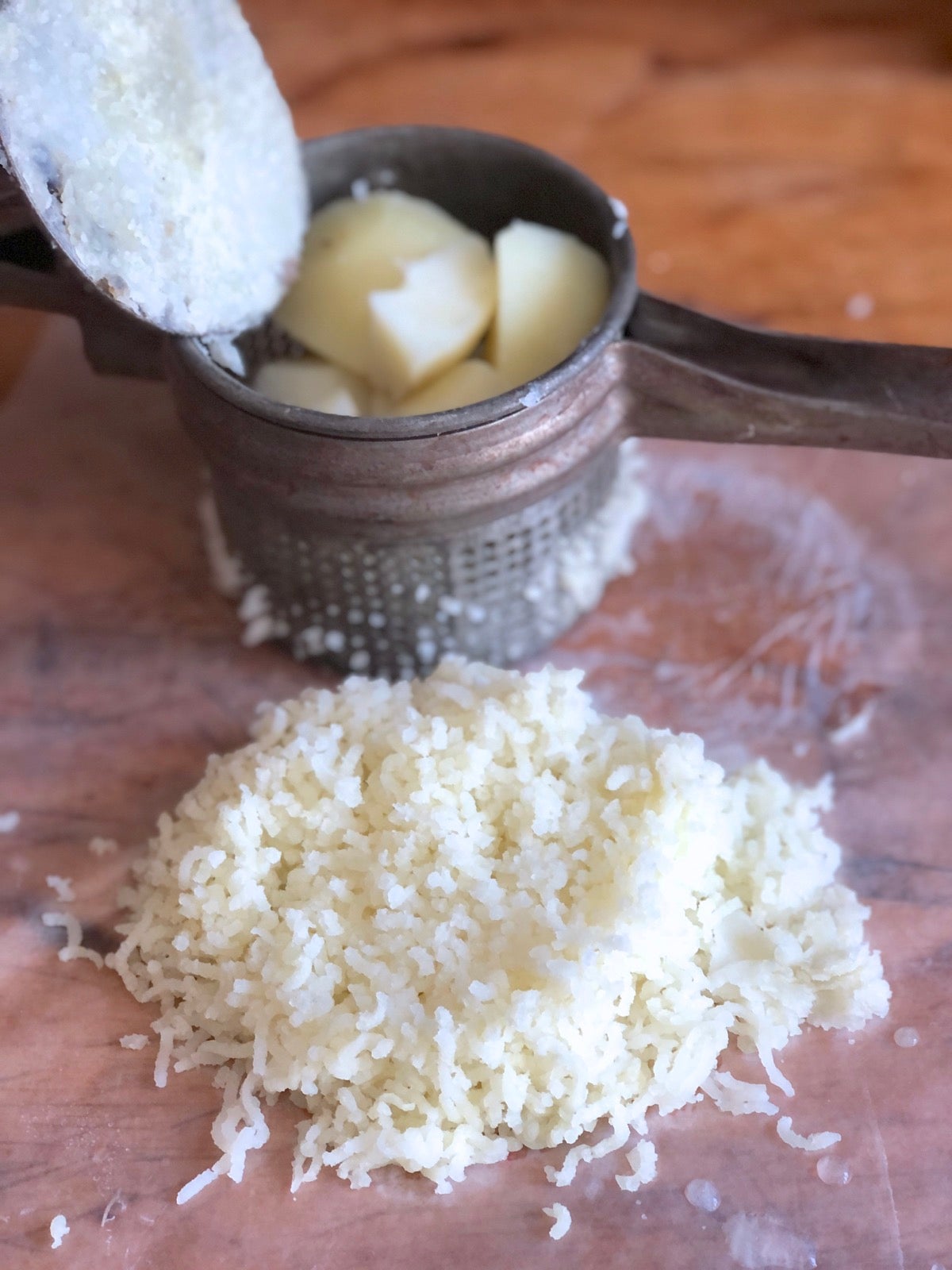 Mound of riced potatoes on a piece of waxed paper, potato ricer in the background. put through a ricer