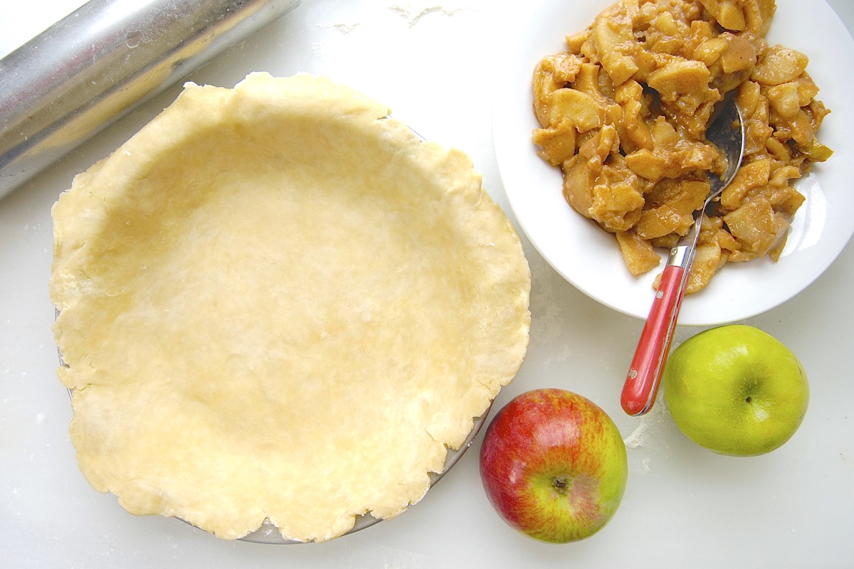 Rolled out pie crust in a pan alongside fresh apples and a bowl of apple pie filling.