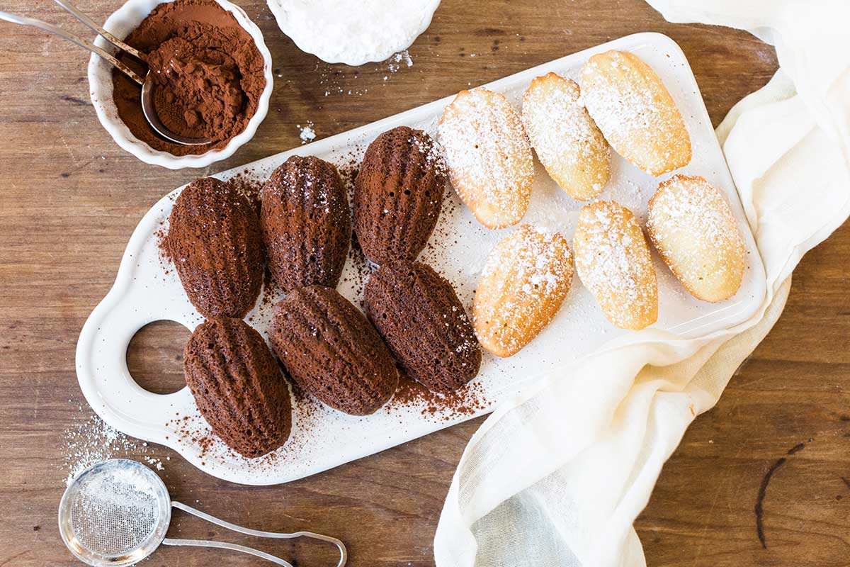 A platter of chocolate and vanilla madeleines next to small bowls of cocoa powder and confectioner's sugar