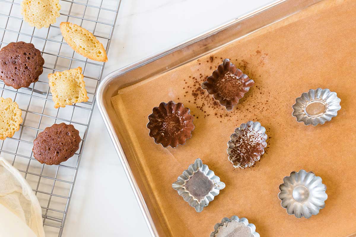 Mini tart shells greased and dusted with cocoa powder next to a few mini madeleines on a cooling rack