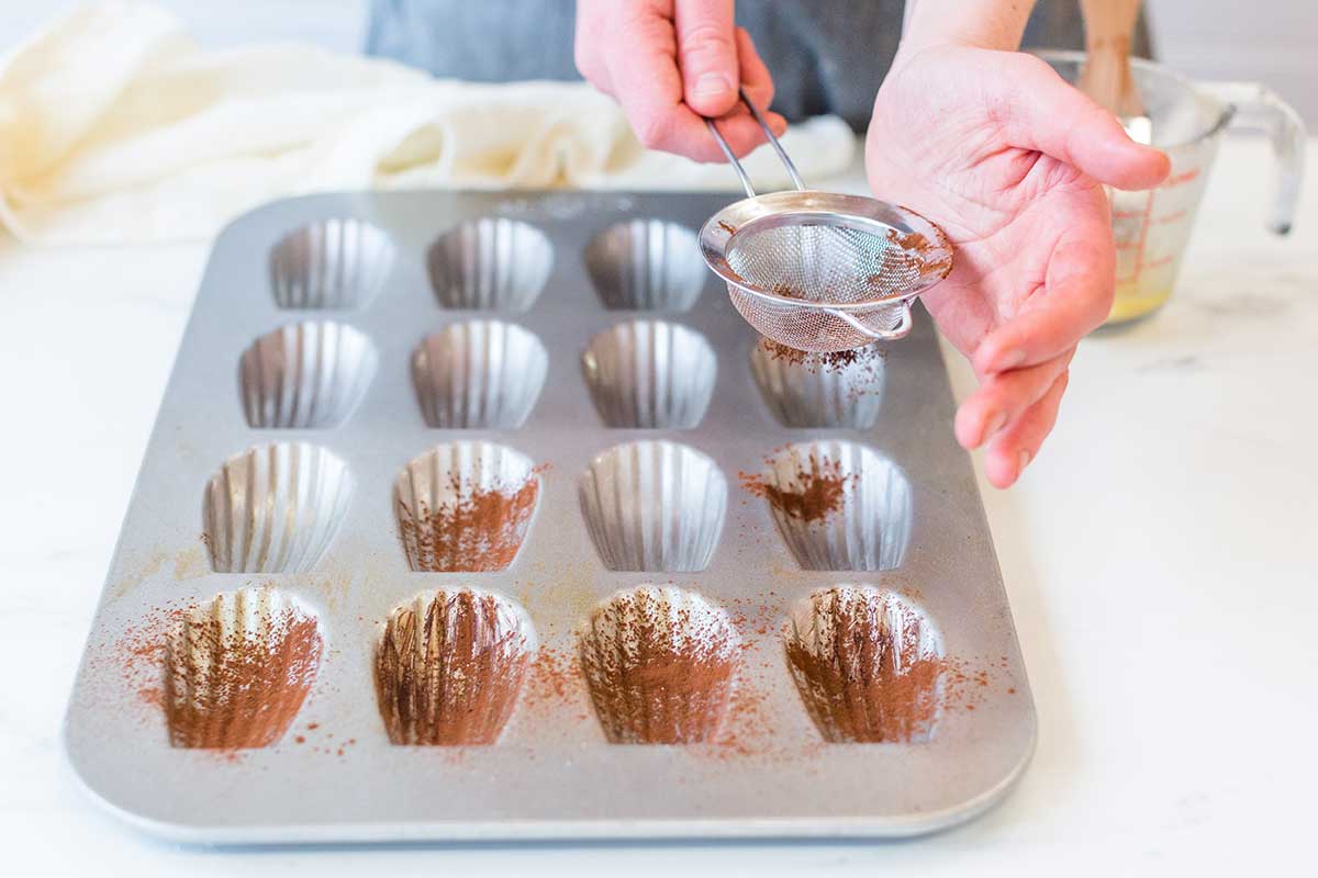 A baker preparing a madeleine pan by dusting each well with cocoa powder