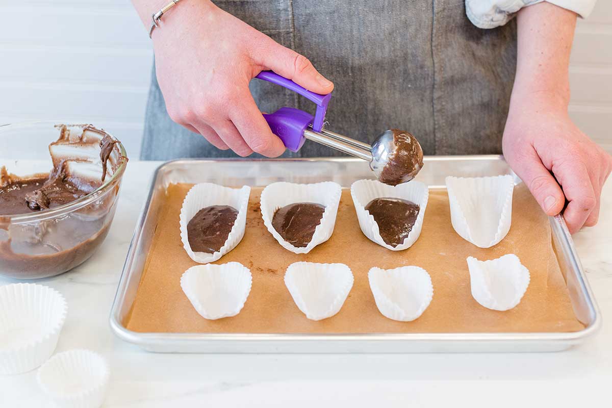 A baker scooping chocolate madeleine batter into free-standing cupcake papers, opened to look like ovals