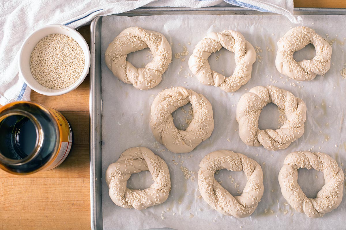Twisted Turkish bagel dough, unbaked, tipped in molasses bath and topped with sesame seeds on a baking sheet
