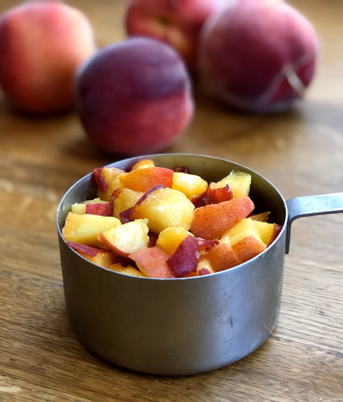 Diced unpeeled peaches in a measuring cup, whole unpeeled peaches in the background.