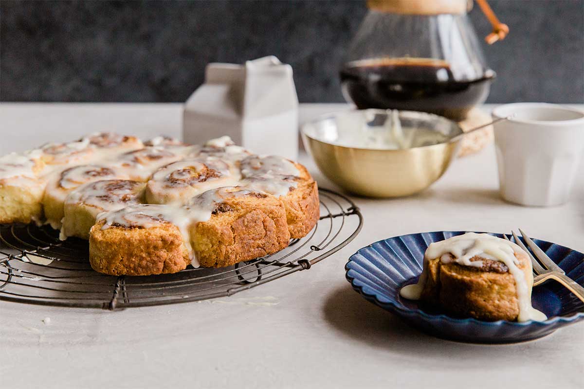 A batch of Instant-Gratification Cinnamon Buns with a few on plates and a coffee carafe in the background