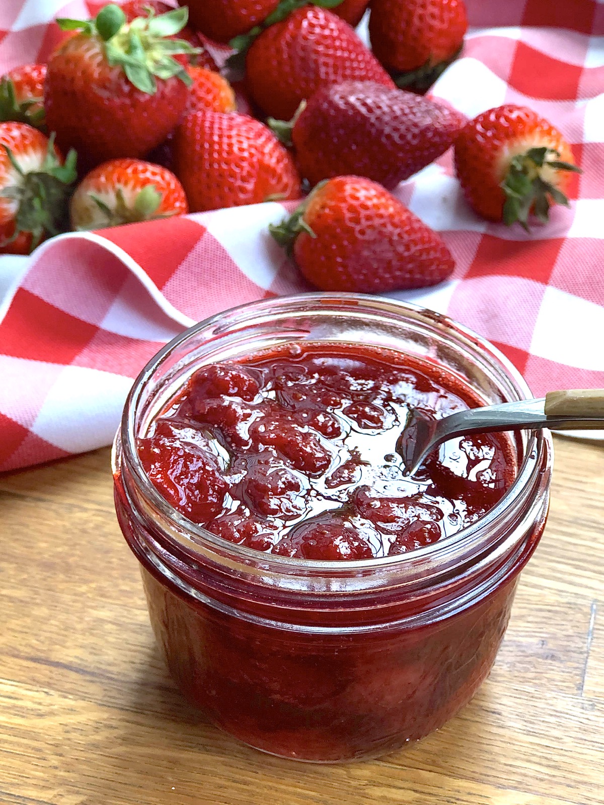Mason jar of strawberry jam with spoon, fresh berries in the background.