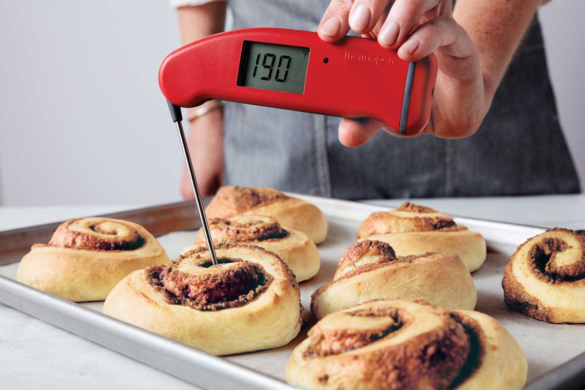 Baked cinnamon rolls on a baking sheet, one's temperature being taken with a probe thermometer and showing 190°F. 