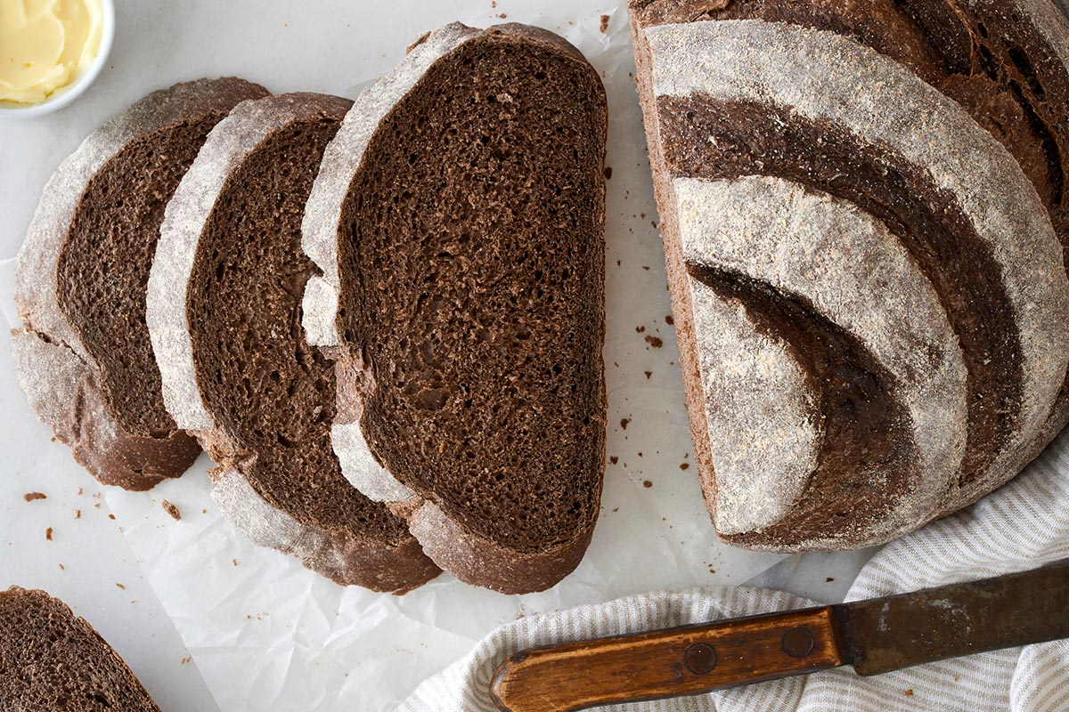 A dark brown loaf of pumpernickel bread shaped into a boule and dusted with flour