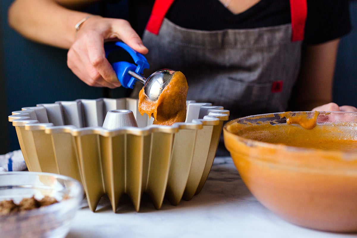 A baker using a muffin scoop to portion pumpkin cake batter into a Bundt pan