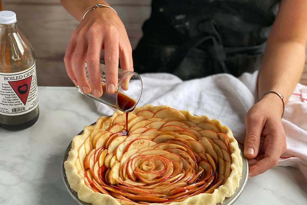 A baker drizzling boiled cider over an unbaked rose apple pie
