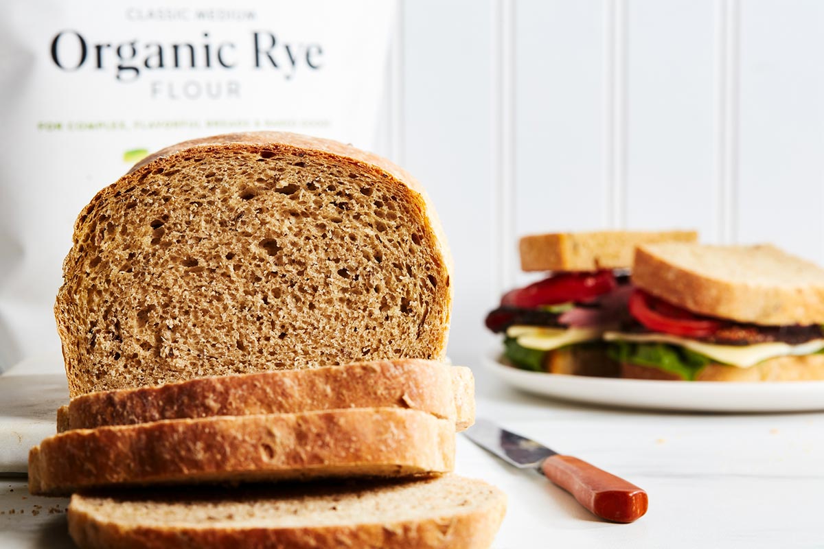 A loaf of rye sandwich bread with a bag of organic medium rye flour in the background