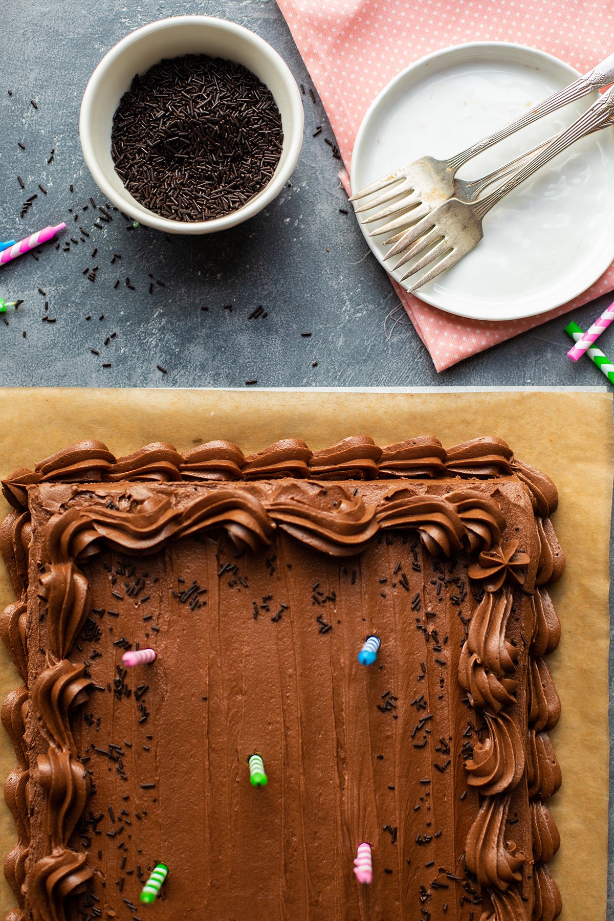 A Classic Birthday Cake topped with birthday candles and a bowl of chocolate sprinkles and plates off to the side