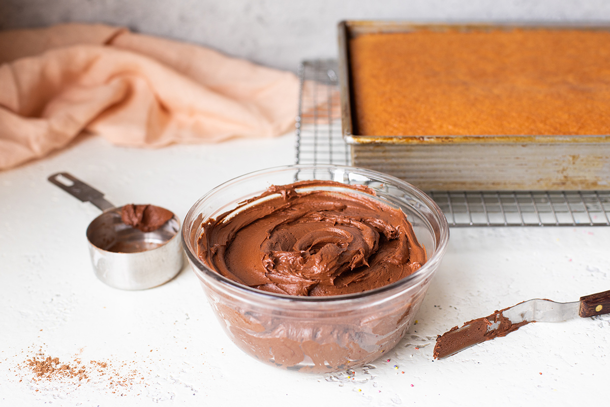 A bowl of chocolate frosting with a spatula off to the side and an unfrosted cake in the background