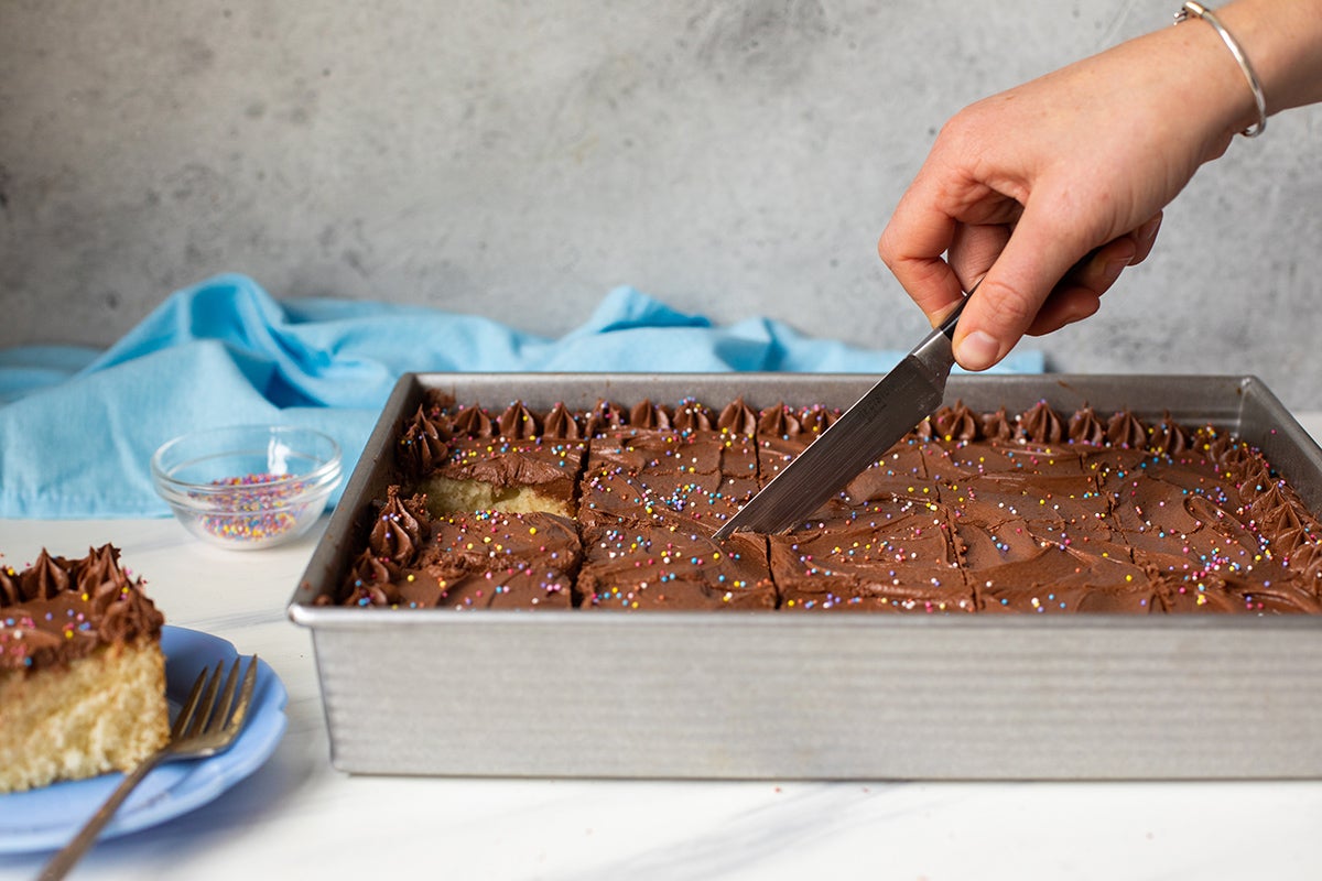 A baker slicing a 9" by 13" sheet cake with chocolate frosting into pieces